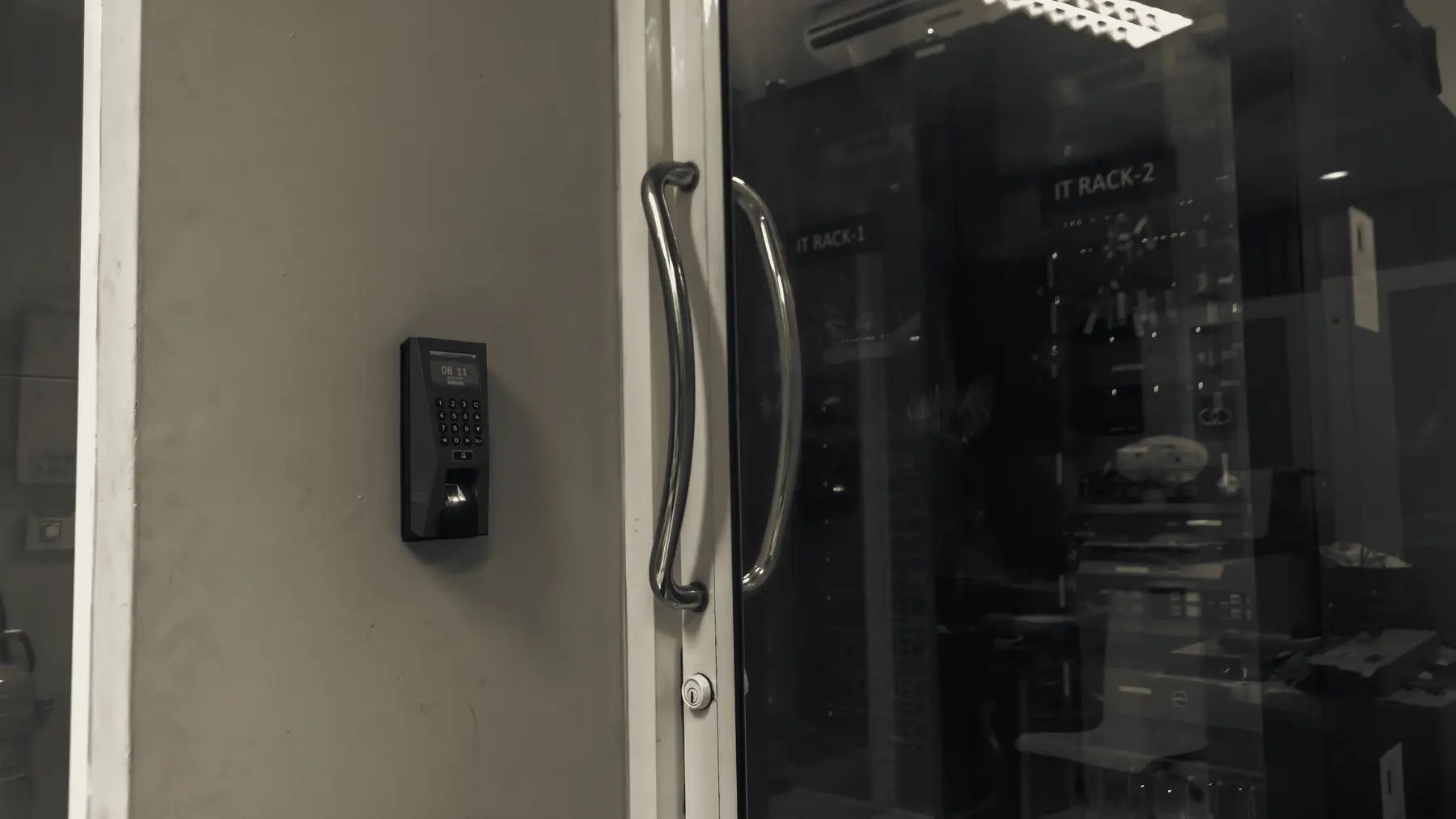 A fingerprint and keypad reader mounted beside the glass door of a server room containing IT racks.